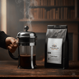 French press with steaming coffee next to a bag of Two Mugs French Roast Coffee on a wooden surface.