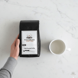 Overhead product photo for scale, showing the Two Mugs Coffee product bag next to a standard 12oz ceramic mug on a light kitchen counter to illustrate size and quantity.